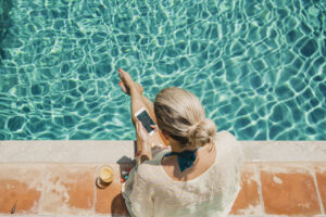 Vogelperspektive auf eine blonde Frau die am Pool sitzt und auf ihr Handy schaut.